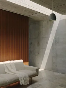 A minimalist living space featuring a concrete wall and wooden paneling with a gray pendant light and beige sofa with a blank