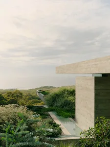 Modern minimalist house with concrete walls and large glass windows overlooking greenery and the ocean.