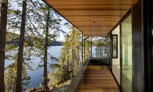 Modern house with glass wall and deck overlooking a forest and lake.