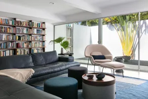 A modern dark living room with a black leather sofa, a beige armchair, a bookshelf, and greenery outdoors.