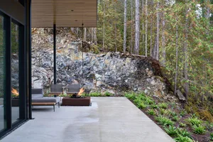 Concrete outdoor patio with industrial metal fire pit and wood furniture amongst tall pine trees