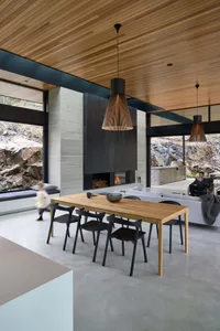 A modern, open-plan living and dining room featuring a wood dining table, concrete fireplace, and contemporary pendant lights