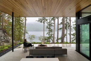 Modern living room with concrete furniture, glass walls, and a fireplace, overlooking a forest and lake.