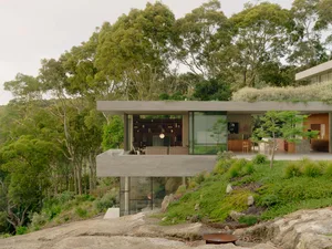 A modern minimalist house with concrete and glass features set in a natural, forested landscape.