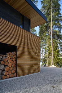 An exterior view of a modern residential building featuring wooden slats, surrounded by trees and a gravel driveway.