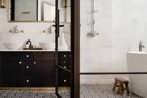 A modern residential bathroom featuring a white marble floor, white marble walls, and a black vanity with white sinks.