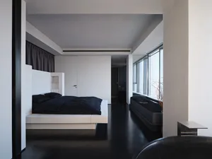 A minimalist bedroom with a white concrete bed platform, black bedding, and a large window.
