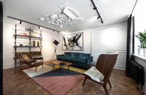 A modern minimalist living room featuring a blue velvet sofa, wooden chairs, and a geometric rug.