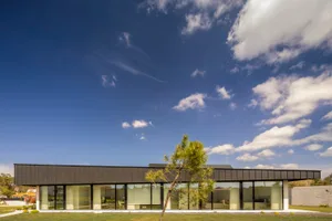 Modern residential exterior with black metal roof and glass windows, set against a clear blue sky with white clouds.