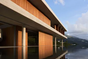Modern architectural building with concrete structure and wooden accents reflected in a water body, set against a mountainous