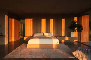 A minimalist bedroom featuring a bed on a wooden platform, surrounded by wooden walls and floor, with sunlight filtering thro
