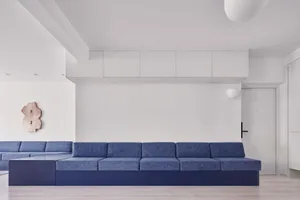 A modern minimalist living room featuring a blue sofa with a dark blue base and a rectangular metallic wall ornament.