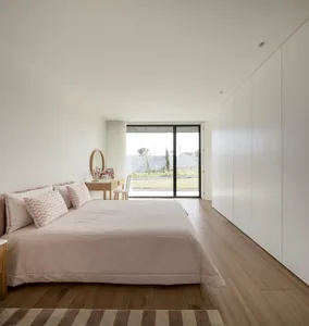 Modern minimalist bedroom with white walls, wooden flooring, and a large window overlooking a garden.