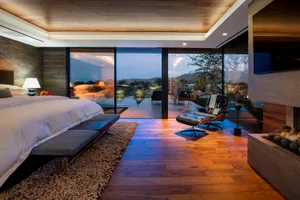 Modern bedroom featuring brown wood flooring, large windows with outdoor view, and a cozy seating area.
