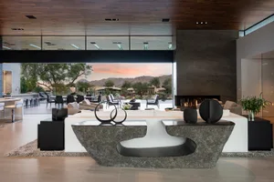 A modern living room with concrete furniture, wood accents, and a large glass window overlooking a desert landscape.