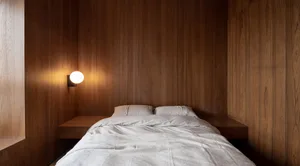A minimalist bedroom with wooden walls, a white bedspread, and a single wall-mounted light.