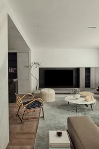 A minimalist living room with woven armchair, wooden sofa, fur pouf, and coffee table on a grey rug.