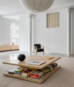 A modern living room featuring white walls, a large wooden coffee table stacked with books and a black decorative object, a b