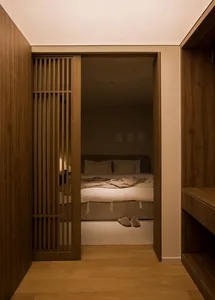 A modern minimalist bedroom with wooden walls and floor, featuring a bed with white bedding and wooden slatted headboard.