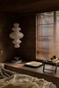 A minimalist bedroom featuring wooden accents, a window with blinds, and a lamp with a geometric design.