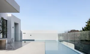 A modern minimalist house exterior with a swimming pool terrace featuring glass railings, concrete patio, and greenery