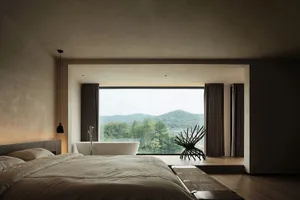 A minimalist bedroom with a concrete floor, a large window overlooking nature, and a comfortable bed.