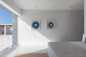 A minimalist bedroom with a concrete floor, featuring a bed with a white mattress and a blue wall with abstract circular artw