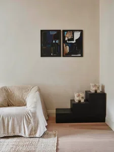 A minimalist living room with a white couch, abstract artwork, and black geometric side table on a wooden floor.