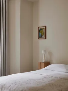 A minimalist bedroom features grey drapery, a wooden bed frame, and a marble lamp on a wooden nightstand.