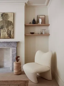 A cozy corner of a minimalist residential interior featuring a white textured lounge chair, a marble fireplace, wood shelves,