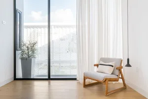 Modern minimalist living room with a wooden armchair, white curtains, and a large window.