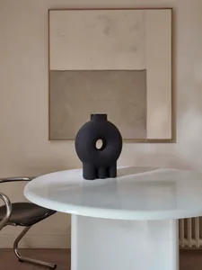 A modern black ceramic vase with a round shape and a central circular cutout sits on a white table.