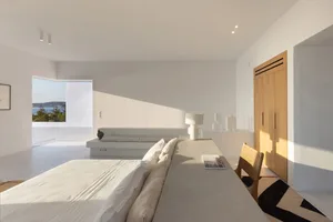 Modern minimalist bedroom with concrete walls and furniture, featuring a large bed and a desk area with a view of the outdoor