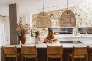 Two children sitting on a kitchen island with wooden chairs, decorated with dried flowers in a vase and large woven pendant l