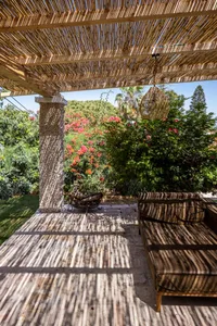 Outdoor wooden pergola with wicker furniture and cement bench surrounded by lush greenery and blooming flowers.