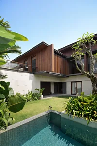 A modern residential pool house with a concrete, wood, and tile exterior, featuring lush landscaping and a swimming pool surr