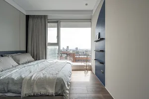 A modern, minimalist bedroom with timber flooring, a large window overlooking a cityscape, and simple furniture.