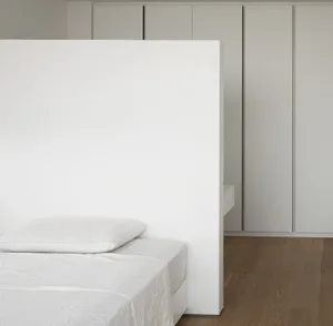 A minimalist bedroom with white walls, a white bed, and wooden flooring.