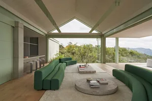 Modern residential interior with green sofas, round coffee table, and glass walls overlooking greenery.