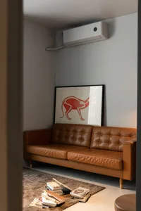 A modern minimalist living room featuring a brown leather couch, a framed red cat skeleton print, and a pile of books on the 