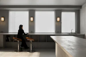 Minimalist kitchen featuring concrete countertops and modern design, with a woman sitting on a stool by the window.