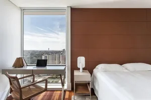 Modern bedroom with a large window overlooking a cityscape, featuring a white bed, white desk, and brown wooden flooring.