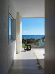 A minimalist residential space with a beach view featuring a wooden chair, glass window, and white fabric rug.