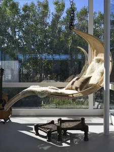 A modern indoor swing with a wicker frame and plush cushions hanging from a ceiling hook, surrounded by large windows with a 