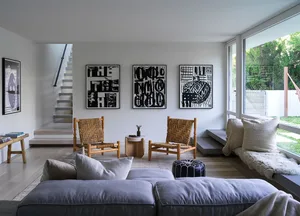 A modern living room with a white minimalist aesthetic, featuring woven-straw armchairs, a sofa with cushions, and abstract b