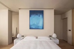 現代簡約臥室，床頭上方懸掛藍色抽象畫作，白色床具，床邊桌上有兩盞圓柱形燈具