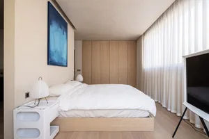 A modern minimalist bedroom with white furniture, a large window, and a blue abstract painting above the bed.