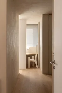 A minimalist residential interior with neutral tones and a small white chair near a window.