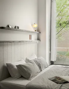 A minimalist bedroom with concrete headboard and grey bedding, featuring a floating concrete shelf with a table lamp and smal