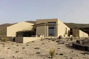 Modern minimalist house exterior with sandstone and concrete materials, featuring a covered patio and desert landscaping.